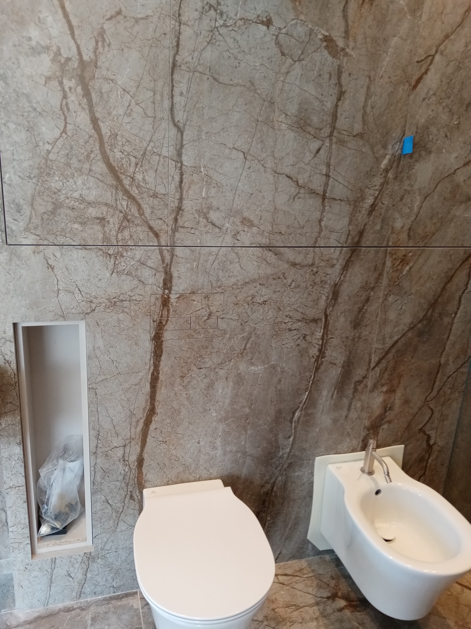 Wykończona łazienka w Pruszkowie z marmurowymi płytami na ścianach, biała toaleta i bidet, wnęka w ścianie z workiem na śmieci.