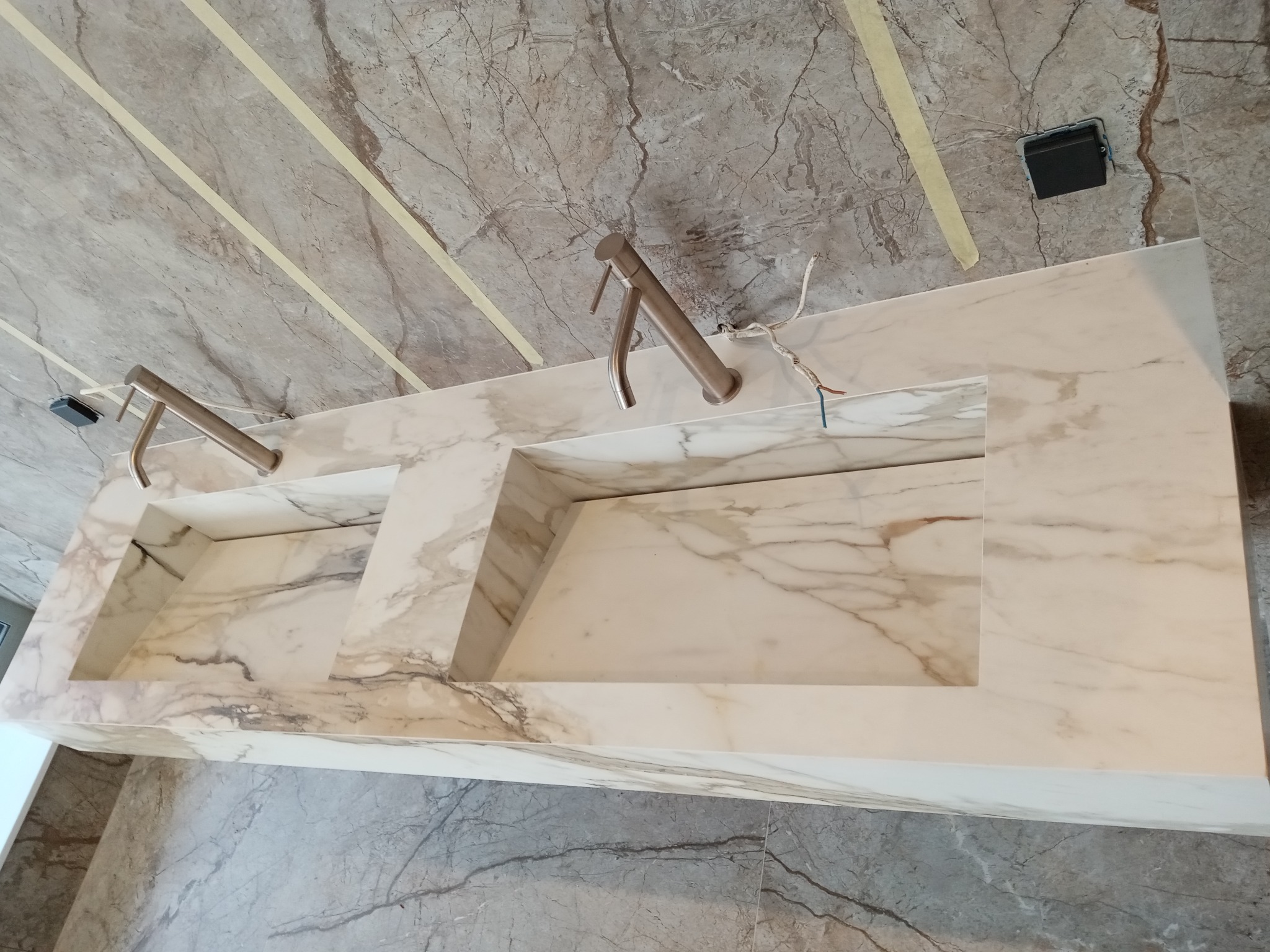 Podwójna umywalka z blatem z marmuru kararyjskiego, zamontowana na tle marmurowych płytek ściennych, z widocznymi metalowymi kranami i elementami instalacji elektrycznej.