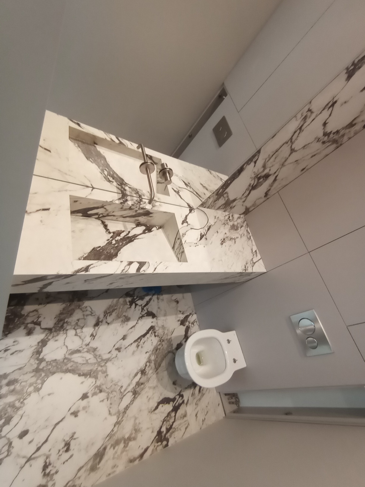 Nowoczesna łazienka z marmurową umywalką wpuszczaną w blat i toaletą podwieszaną, wykończona płytkami w odcieniach bieli i szarości.