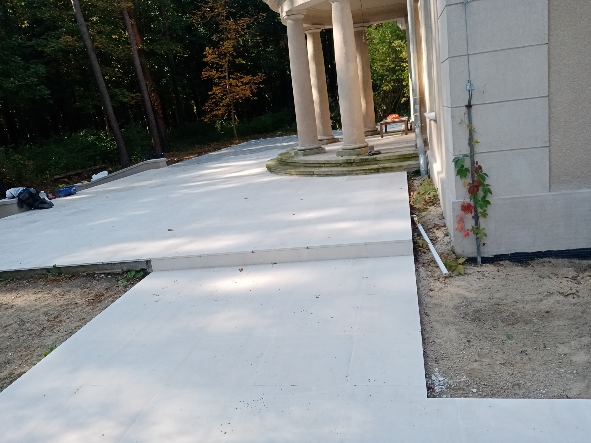 Jasny, wyłożony płytkami taras z kolumnadą w tle, częściowo w cieniu drzew, z widocznymi śladami prac wykończeniowych i roślinnością przy ścianie budynku.