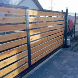 Przesuwna brama wjazdowa z poziomych desek z widocznym automatem do bramy, obok ogrodzenie panelowe i kostka brukowa.