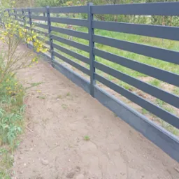 Nowoczesne, poziome ogrodzenie panelowe w kolorze grafitowym, osadzone na betonowej podmurówce. W tle zieleń, z przodu kwitnący krzew. Ujęcie z bliska.