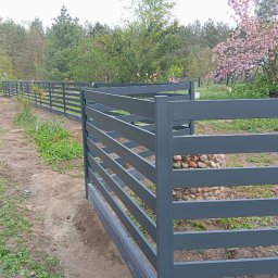 Wojciech Krzyśków Budmak Budowa i Remont - Szare, poziome ogrodzenie panelowe w ogrodzie, z widocznym podmurówką i drzewami w tle. Elementy mocujące słupki ogrodzenia z kamienia.