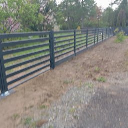 Wojciech Krzyśków Budmak Budowa i Remont - Szary, poziomy płot panelowy z metalu, zamontowany wzdłuż ścieżki. Widoczne słupki i poziome panele. Trawa i drzewa w tle, częściowo zamazane.