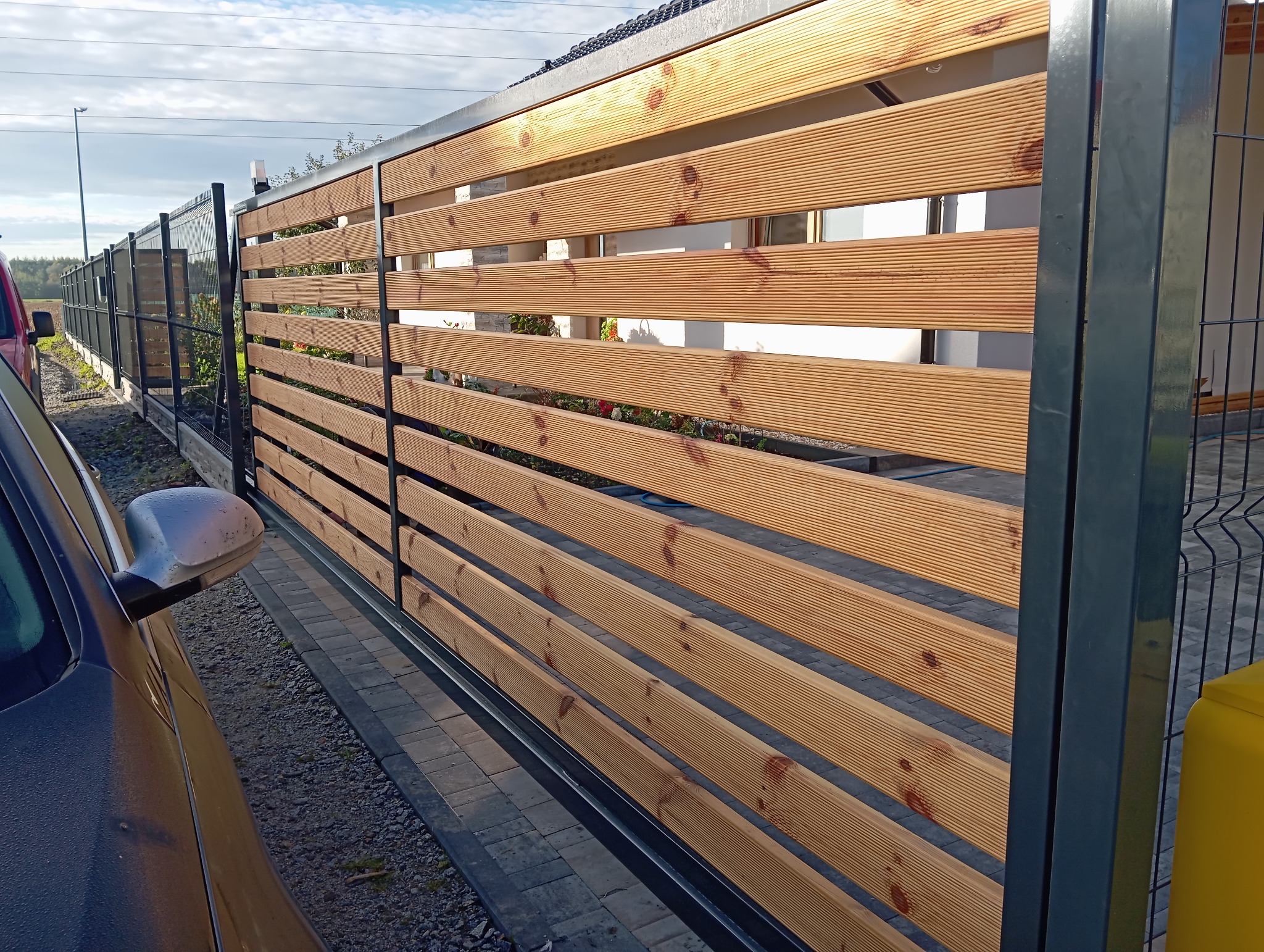 Przesuwna brama wjazdowa z poziomych desek drewnianych w metalowej ramie, widok z boku, z elementami ogrodzenia panelowego w tle.