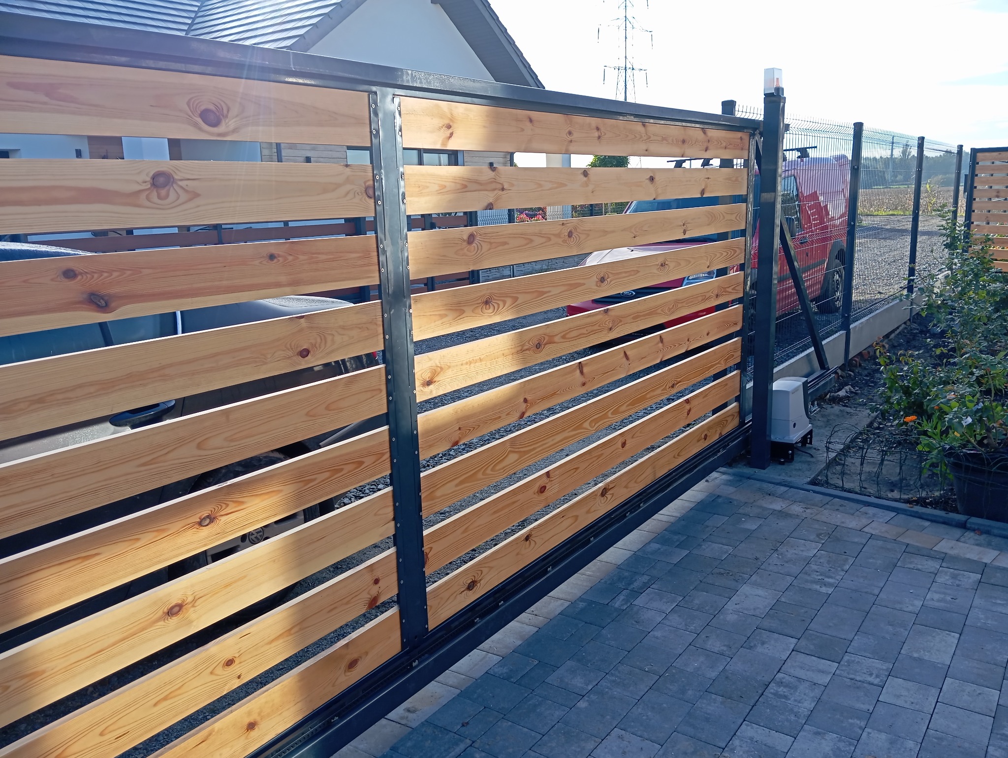 Przesuwna brama wjazdowa z poziomych desek z widocznym automatem do bramy, obok ogrodzenie panelowe i kostka brukowa.