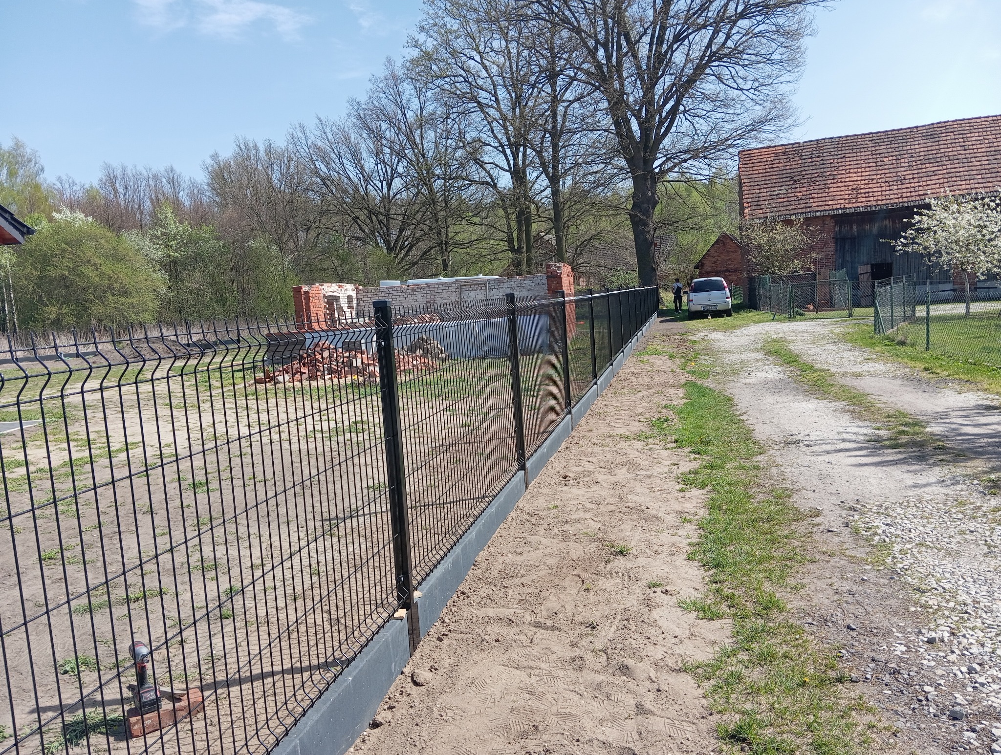 Nowo zamontowane czarne ogrodzenie panelowe z podmurówką, widoczne na tle wiejskiego krajobrazu z budynkami gospodarczymi i stertą cegieł.
