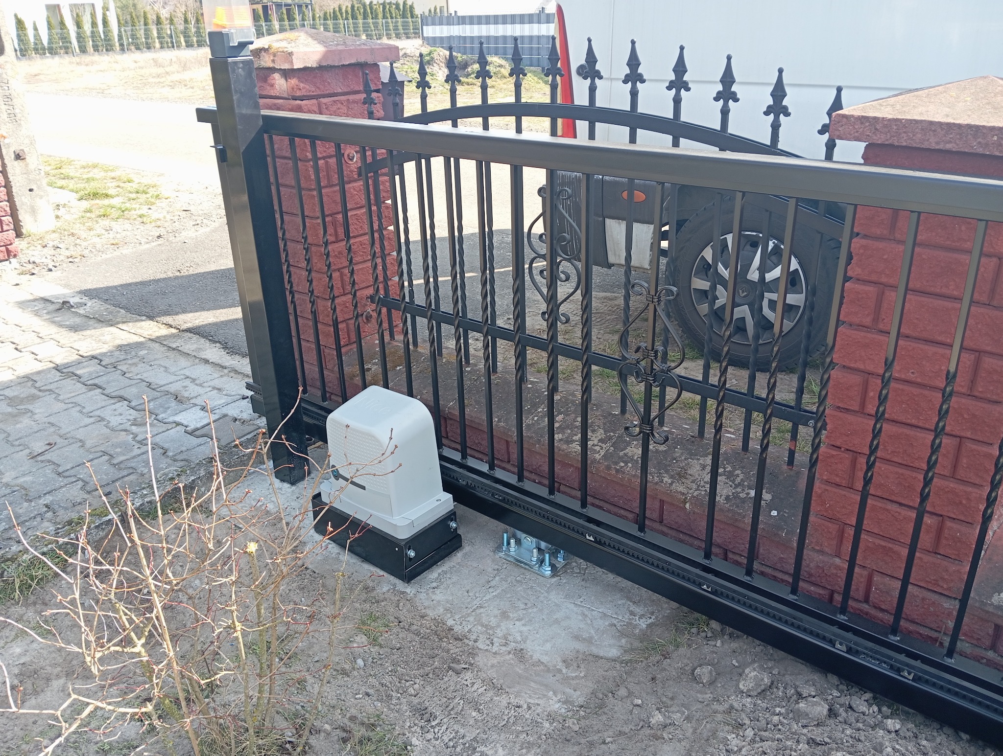 Czarna, metalowa brama przesuwna z dekoracyjnymi elementami, zamontowana na ceglanym słupku, z widocznym napędem automatycznym i elementami mocującymi. W tle widoczny samochód dostawczy.