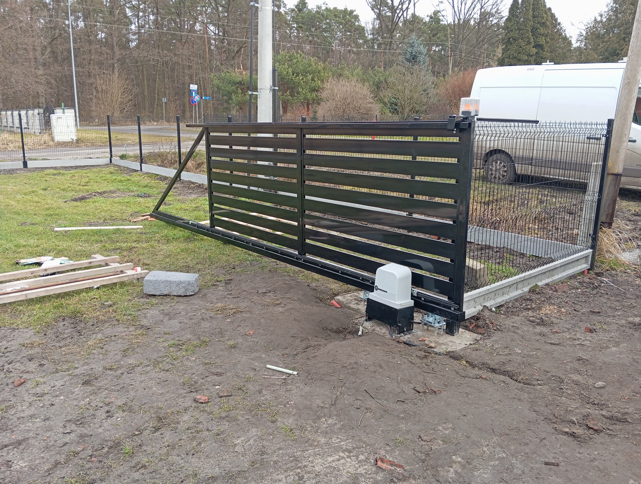 Częściowo zamontowana, przesuwna brama wjazdowa z automatyką, widoczny silnik i ogrodzenie panelowe w tle, teren po pracach montażowych.