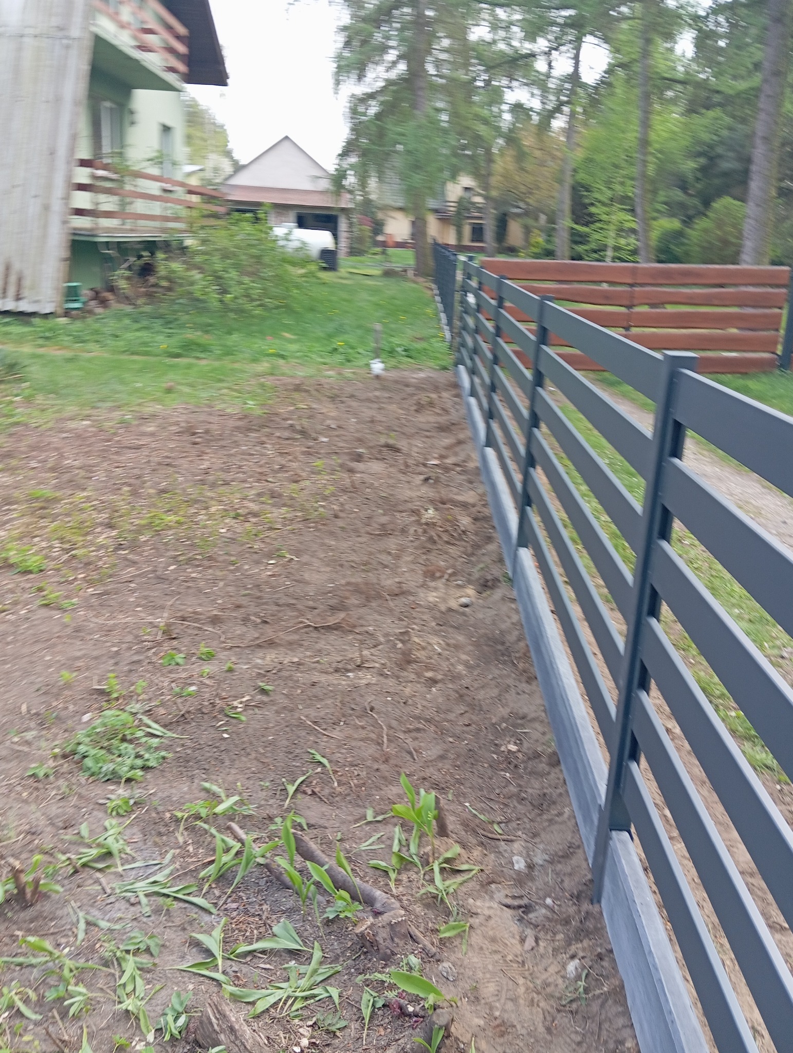 Szary metalowy płot panelowy z betonową podmurówką, widziany z perspektywy gruntu, z domem i drzewami w tle. Widoczne elementy montażowe.