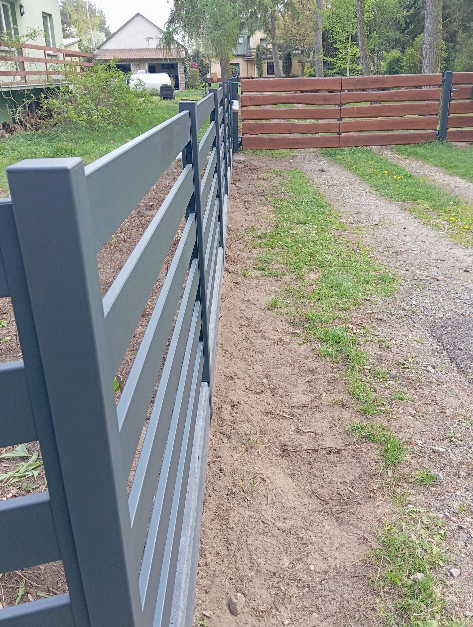 Szary panel ogrodzeniowy z betonową podmurówką, widok z boku, w tle brama z drewnianych desek. Ujęcie z perspektywy.