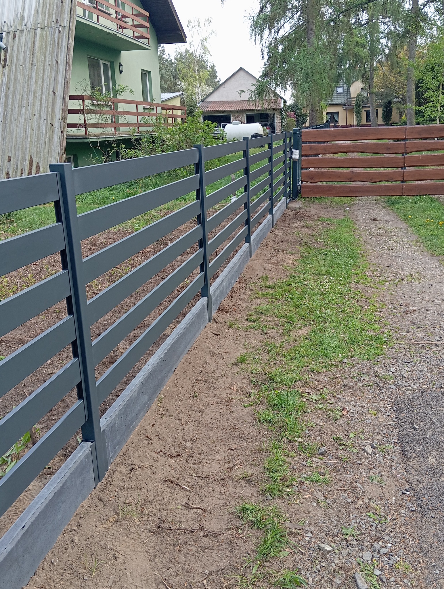 Szare ogrodzenie panelowe z betonową podmurówką, z drewnianą bramą w tle, na tle zieleni i zabudowań. Widok z boku, lekki kąt.