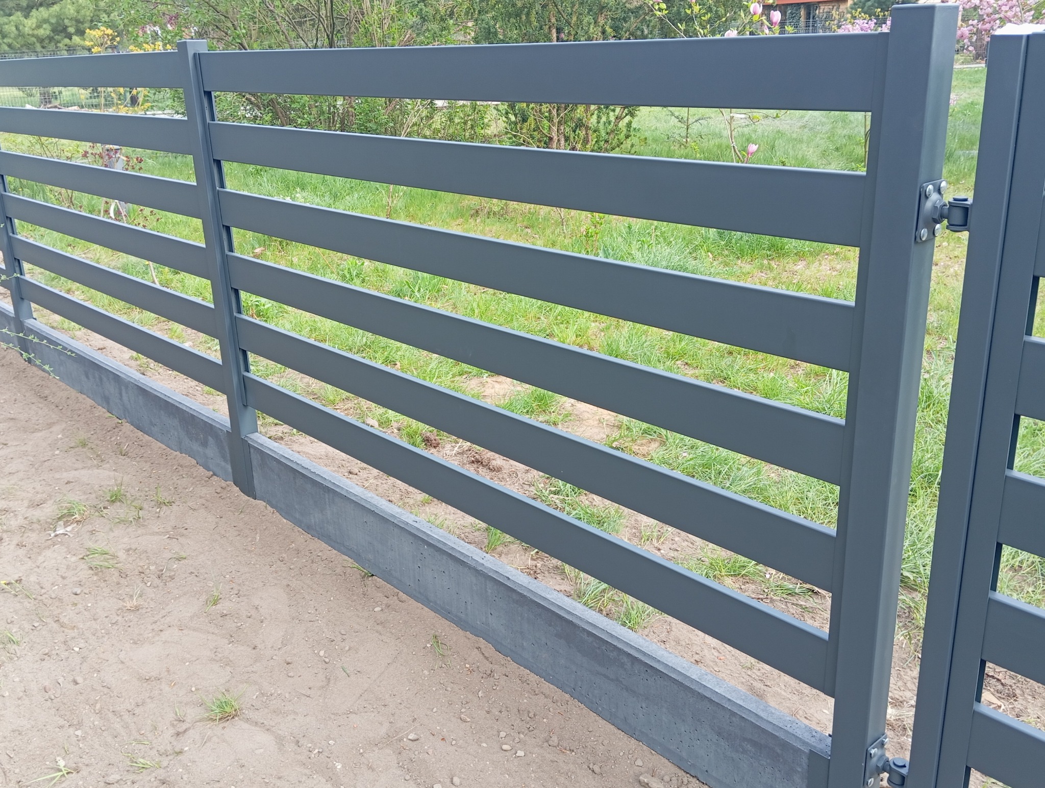 Nowoczesne, szare ogrodzenie panelowe z podmurówką betonową, widok z boku, z elementami mocującymi bramę. Trawa i roślinność w tle.