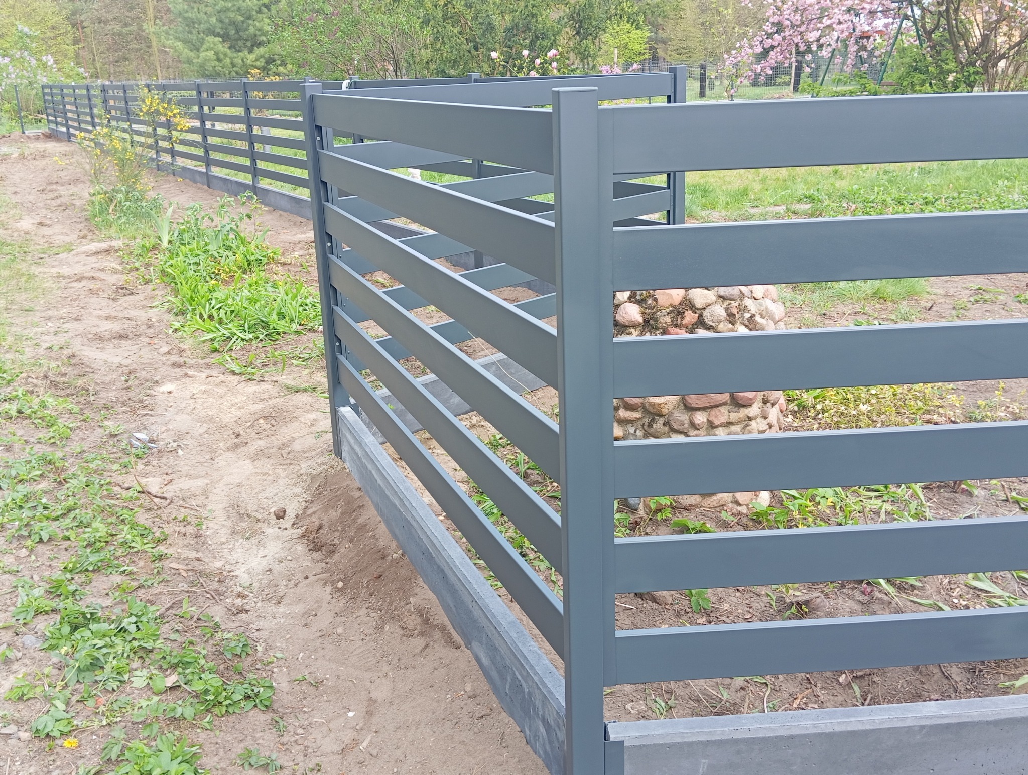 Nowoczesne, poziome ogrodzenie metalowe w kolorze szarym, z betonową podmurówką, na tle zieleni i drzew. Widok z bliska, podkreślający minimalistyczny design.