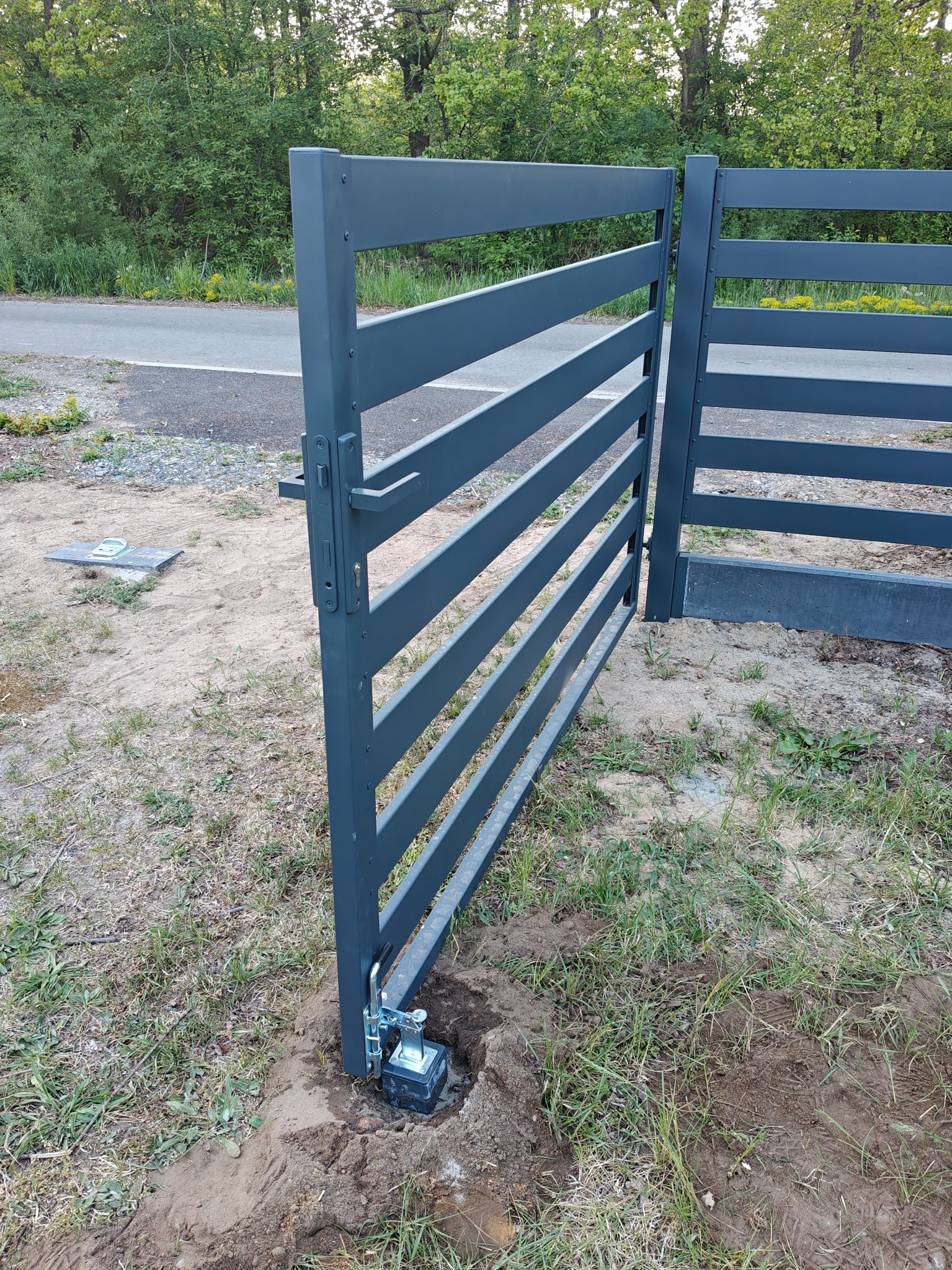 Otwarta, pozioma brama wjazdowa z szarego metalu, z widocznym mechanizmem otwierającym, zamontowana na posesji z zielenią i drogą w tle.