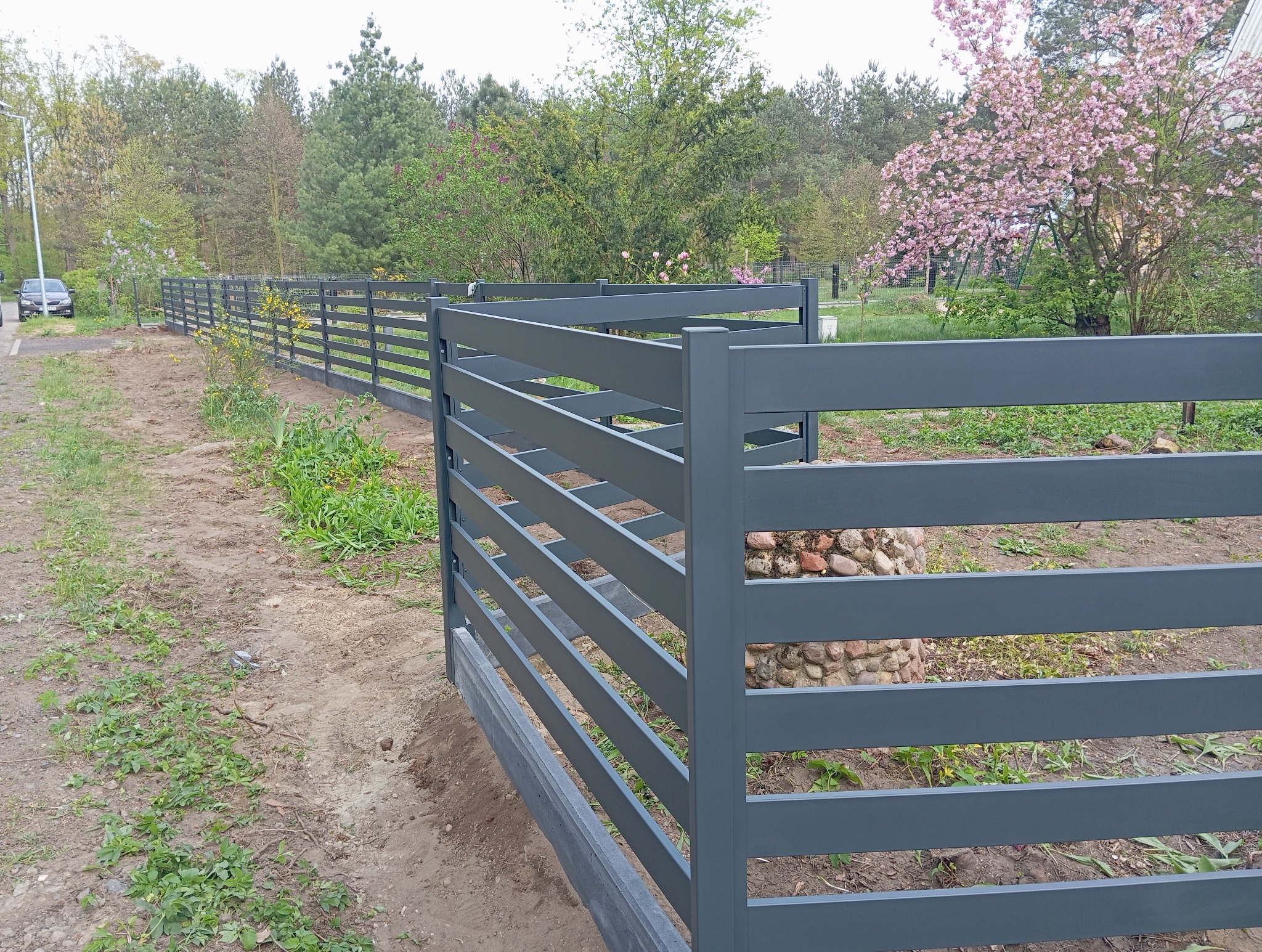 Szare, poziome ogrodzenie panelowe w ogrodzie, z widocznym podmurówką i drzewami w tle. Elementy mocujące słupki ogrodzenia z kamienia.