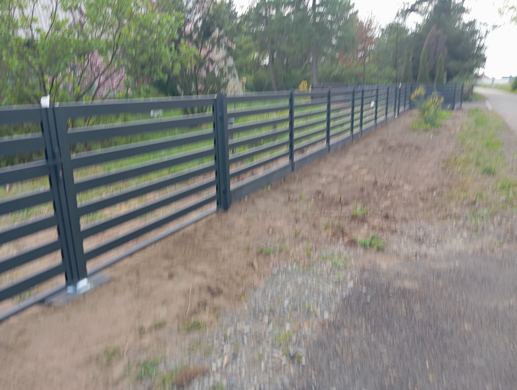 Szary, poziomy płot panelowy z metalu, zamontowany wzdłuż ścieżki. Widoczne słupki i poziome panele. Trawa i drzewa w tle, częściowo zamazane.