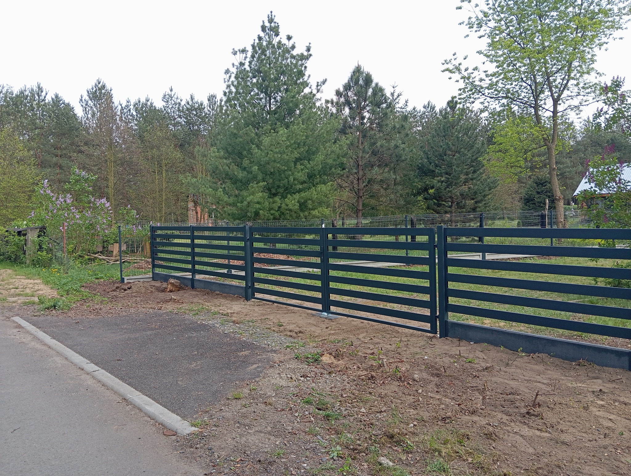 Nowoczesne, poziome ogrodzenie metalowe w kolorze grafitowym, z widoczną bramą wjazdową, na tle zieleni drzew i krzewów. Minimalistyczny design, surowy krajobraz.