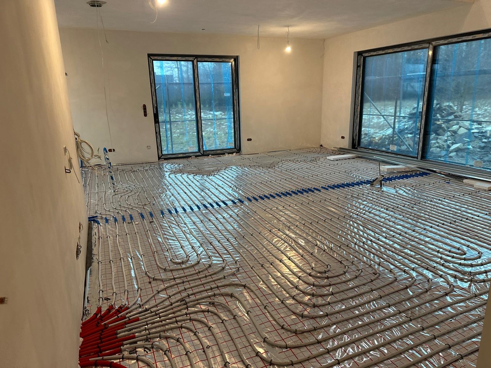 Rozłożona instalacja ogrzewania podłogowego z czerwonymi i niebieskimi złączkami na folii z siatką, widok na duże okna z widokiem na zewnątrz.