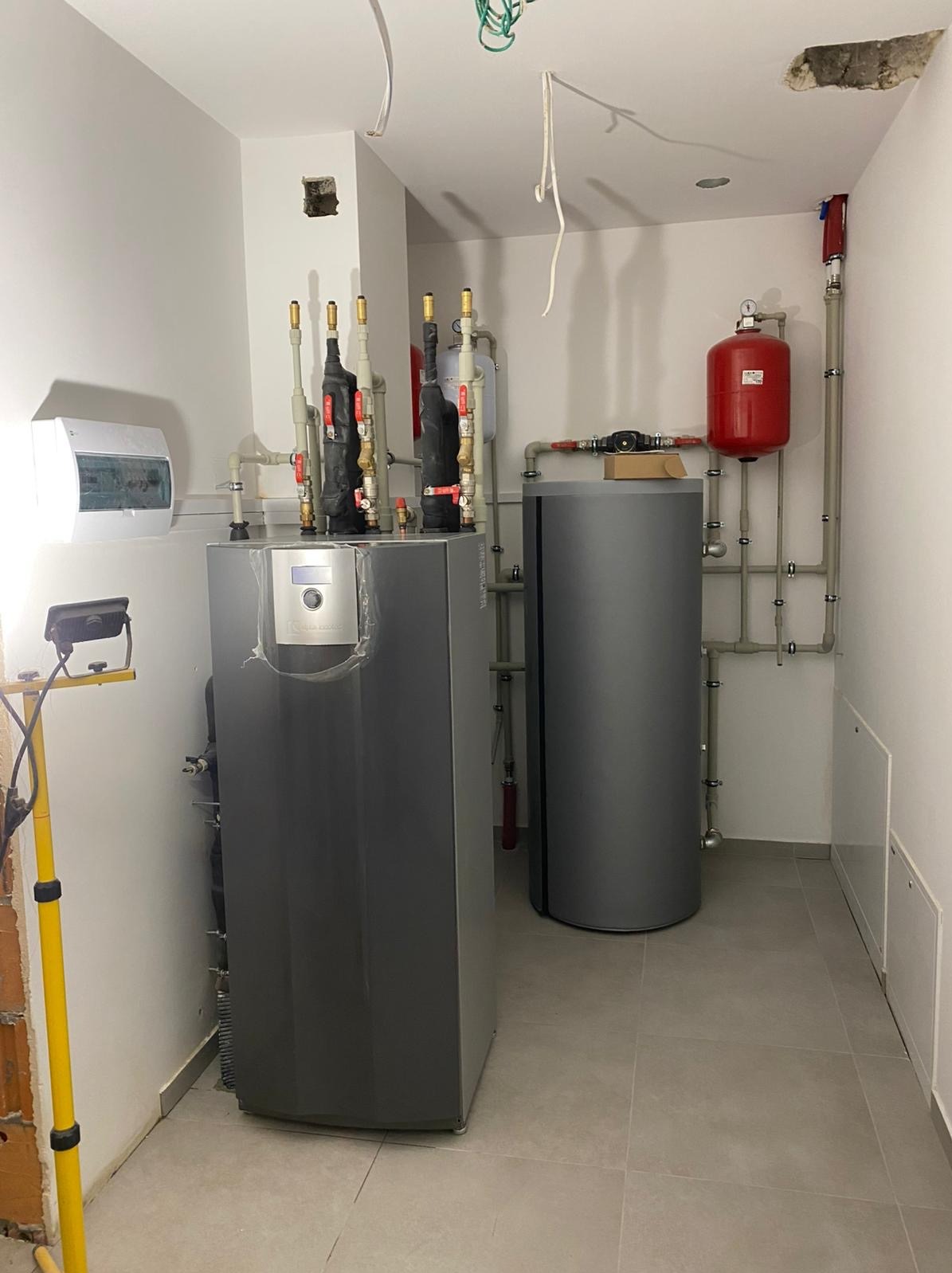 Kotłownia: nowoczesny piec grzewczy w szarej obudowie z widocznymi rurami, zbiornikiem wyrównawczym i instalacją elektryczną, oświetlony lampą roboczą.