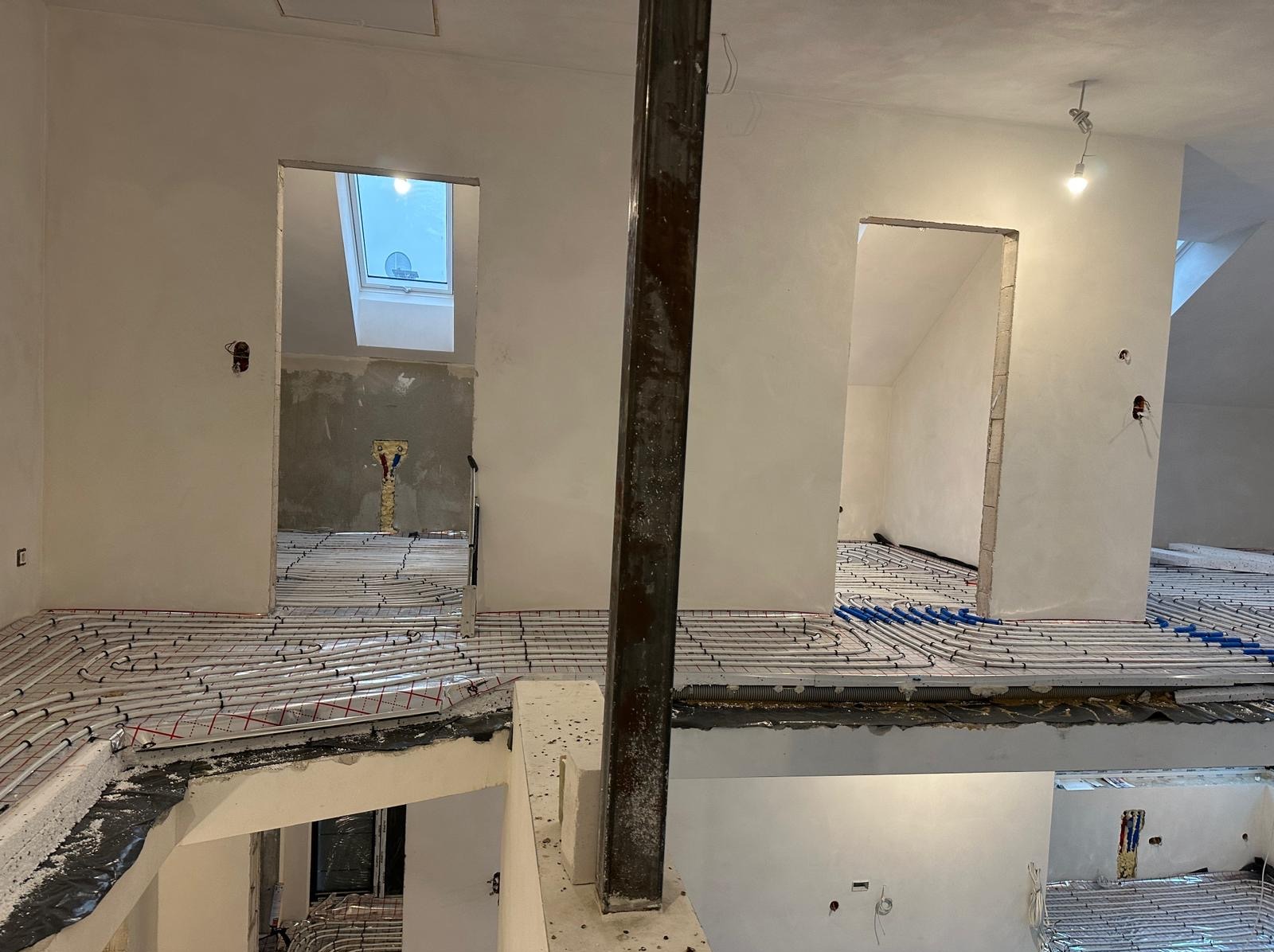 Rozległy widok na wnętrze w stanie surowym z zamontowanym ogrzewaniem podłogowym, widoczne pętle rur w kolorze białym i niebieskim, oraz metalowy słup w centrum kadru.