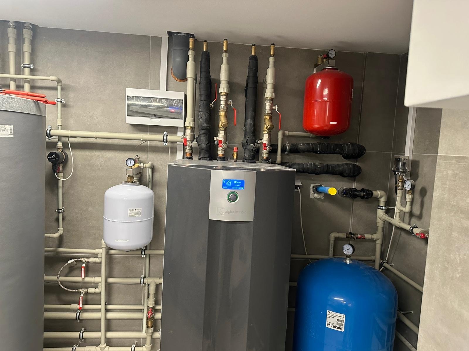 Szara kotłownia z rozbudowaną instalacją hydrauliczną, widoczne rury, zbiorniki wyrównawcze czerwony i niebieski, oraz nowoczesny piec z wyświetlaczem Alpha Innotec.