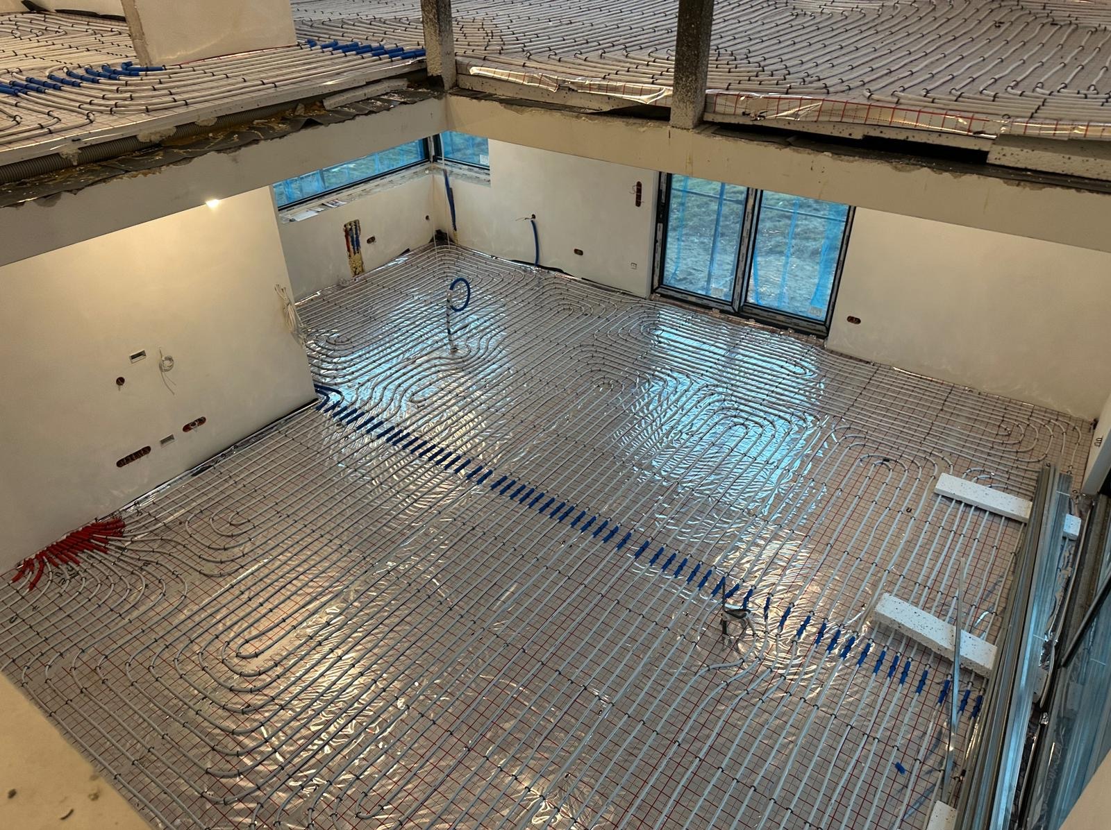 Widok z góry na rozłożone rury ogrzewania podłogowego na folii izolacyjnej, w surowym stanie budynku z widocznymi oknami i elementami konstrukcyjnymi.