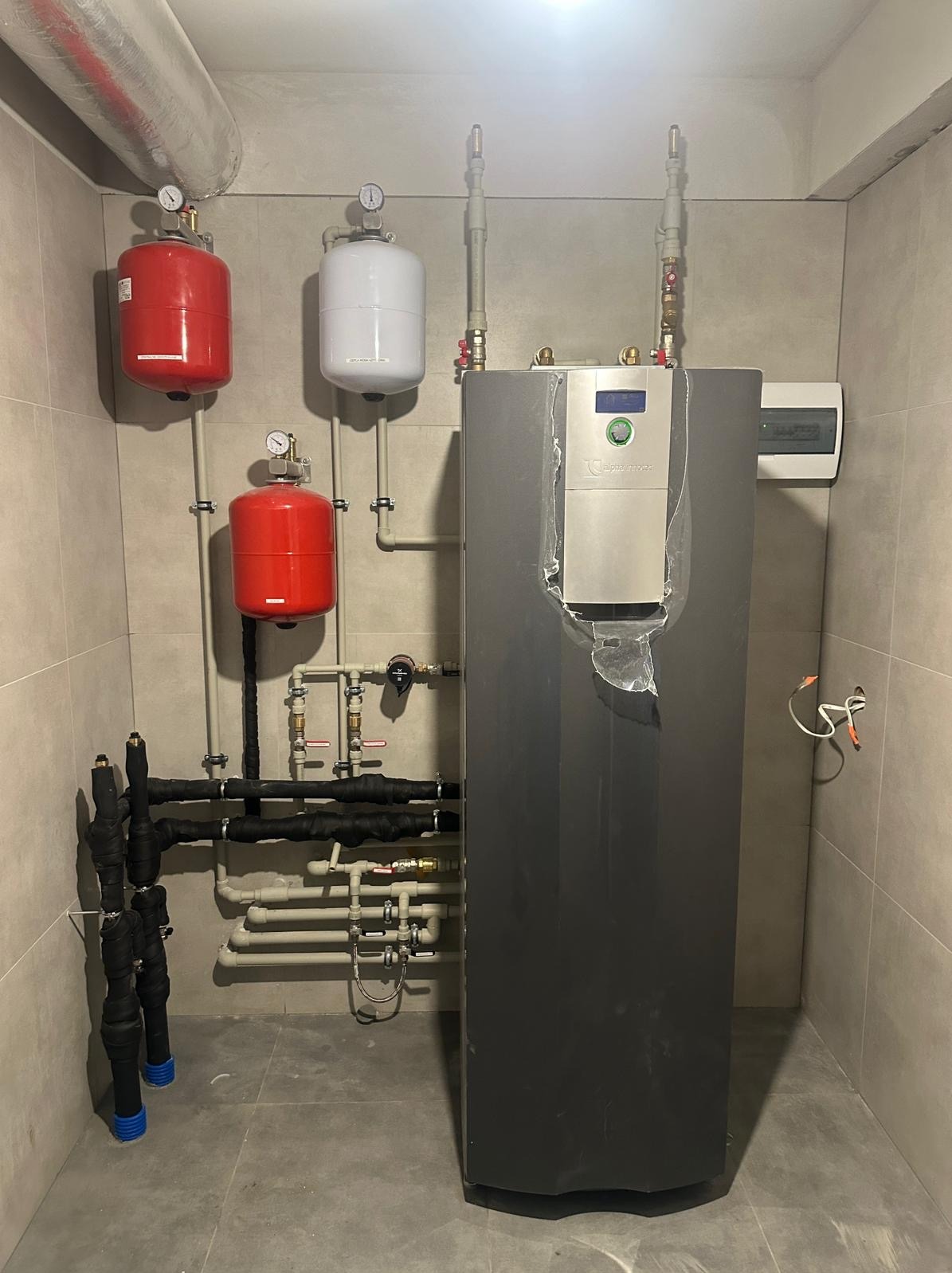 Nowoczesny, kondensacyjny piec grzewczy z trzema zbiornikami wyrównawczymi (dwa czerwone, jeden biały) zainstalowany w pomieszczeniu wyłożonym płytkami ceramicznymi. Widoczne rury grzewcze...