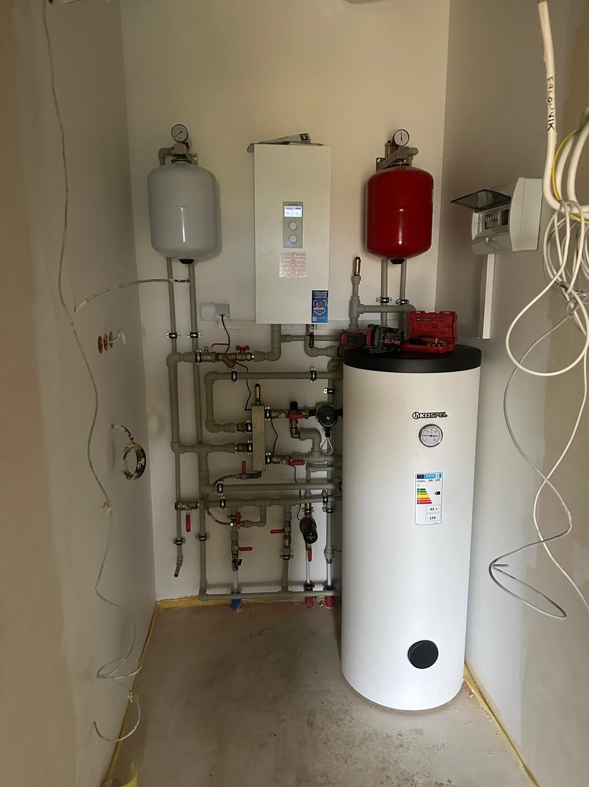 Zestaw instalacji grzewczej w kotłowni: biały zasobnik ciepłej wody Koszel, szare rury z zaworami, czerwony i szary zbiornik wyrównawczy, biały piec z wyświetlaczem, skrzynka elektryczna...