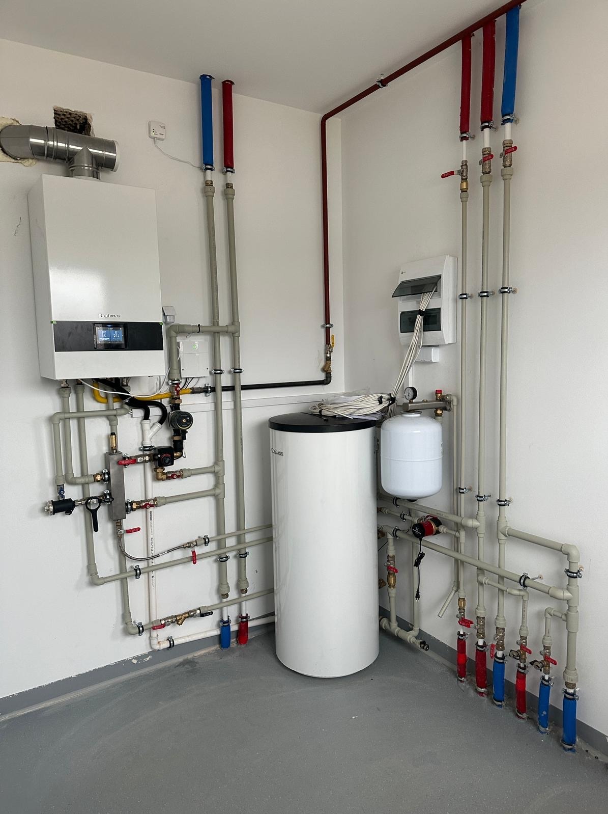 Nowoczesna instalacja grzewcza z piecem gazowym, zasobnikiem ciepłej wody i rozdzielnicą elektryczną w pomieszczeniu technicznym w Ciemnem. Widoczne rury z oznaczeniami kolorystycznymi i liczne...