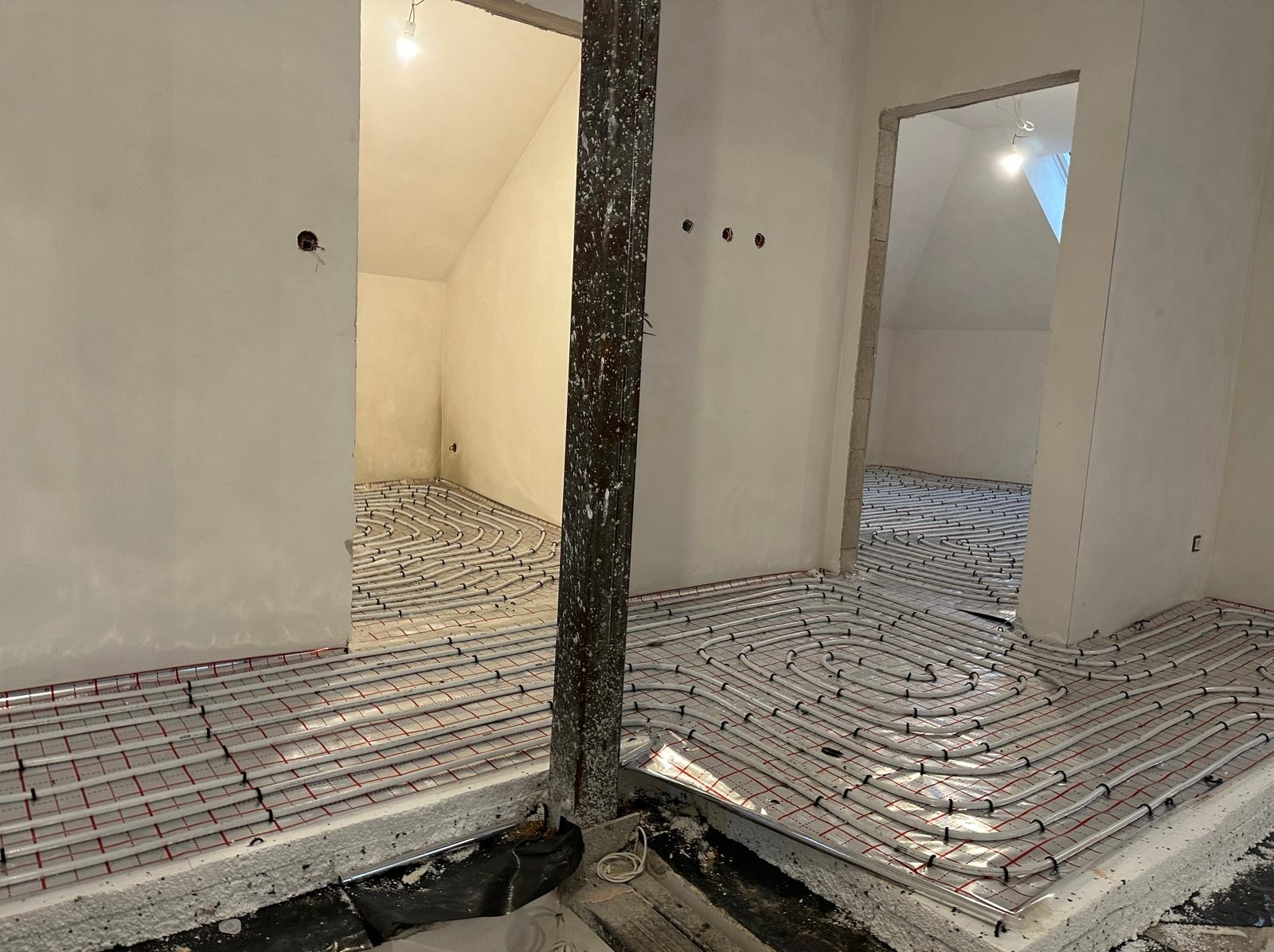 Montaż ogrzewania podłogowego w dwóch pomieszczeniach, rury ułożone na izolacji, widoczny surowy stan ścian i słup konstrukcyjny.