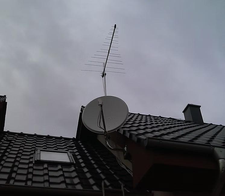 Antena satelitarna i antena telewizyjna zamontowane na dachu pokrytym czarną dachówką, widok z dołu na pochmurne niebo.
