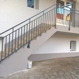 WAWRO Mariola & Mariusz - Zewnętrzne schody betonowe z prostą, metalową poręczą w kolorze grafitowym, prowadzące na mały balkon z metalową balustradą, widok z dołu, obok ściany budynku z małymi oknami.