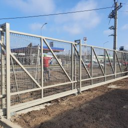 WAWRO - Długa, przesuwna brama przemysłowa z metalowej siatki, pomalowana na jasny kolor, z widocznym mechanizmem przesuwnym i betonową podstawą, na tle stacji benzynowej i słupów energetycznych.