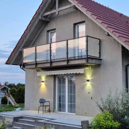 WAWRO - Dom z balkonem z czarną ramą i szklanymi panelami, oświetlony kinkietami, z tarasem i schodami wejściowymi, w tle plac zabaw dla dzieci.