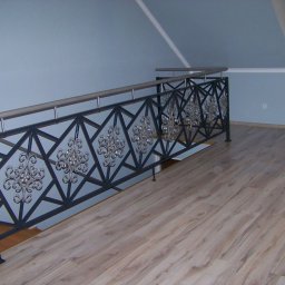 WAWRO - Kuta, czarna balustrada schodowa z ozdobnymi, srebrnymi rozetami i stalową poręczą, widoczna na tle błękitnej ściany i drewnianej podłogi.