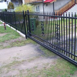 WAWRO - Czarna brama wjazdowa przesuwna z metalowymi prętami i ozdobnymi elementami, widoczna w perspektywie z boku na tle domu z czerwonym dachem i zielenią.