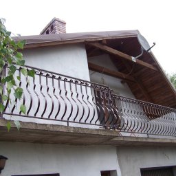 WAWRO - Balkon z metalową balustradą o falistym wzorze, widok z dołu, z fragmentem zwisających gałęzi drzewa w lewym górnym rogu, na tle białej ściany budynku z brązowym dachem i kominem.