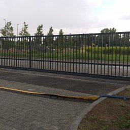 WAWRO - Przesuwna brama przemysłowa z czarnego metalu z zabezpieczeniem najazdowym na kostce brukowej; w tle teren budowy z widoczną koparką.