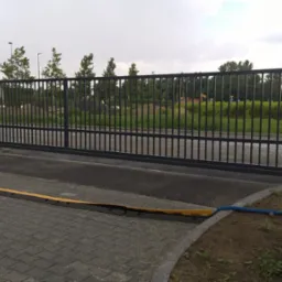 Przesuwna brama przemysłowa z czarnego metalu z zabezpieczeniem najazdowym na kostce brukowej; w tle teren budowy z widoczną koparką.