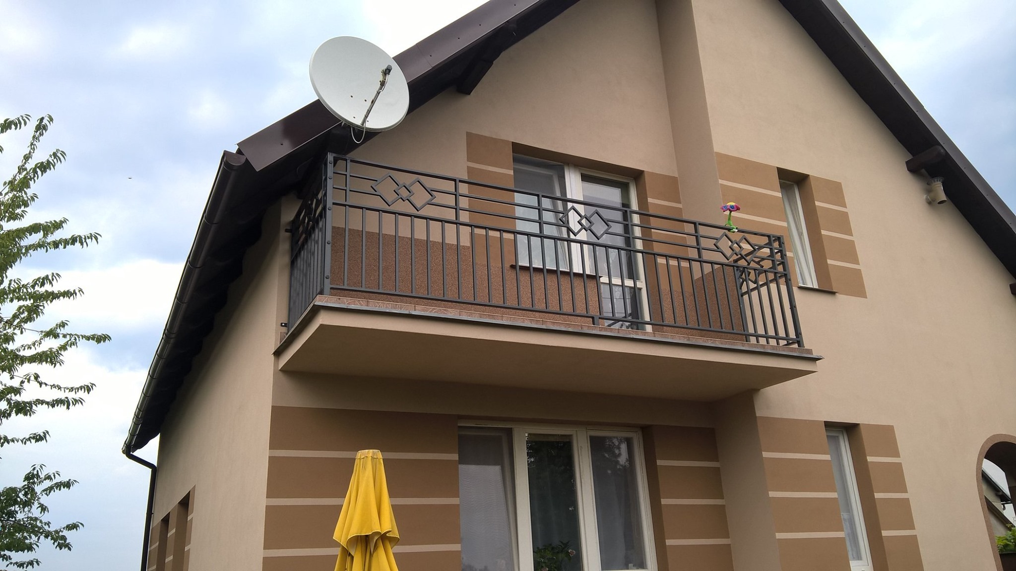 Elewacja domu z balkonem zabezpieczonym czarną, metalową balustradą o geometrycznym wzorze rombów, na balkonie stoi zabawka w kształcie kwiatka, na dachu antena satelitarna.