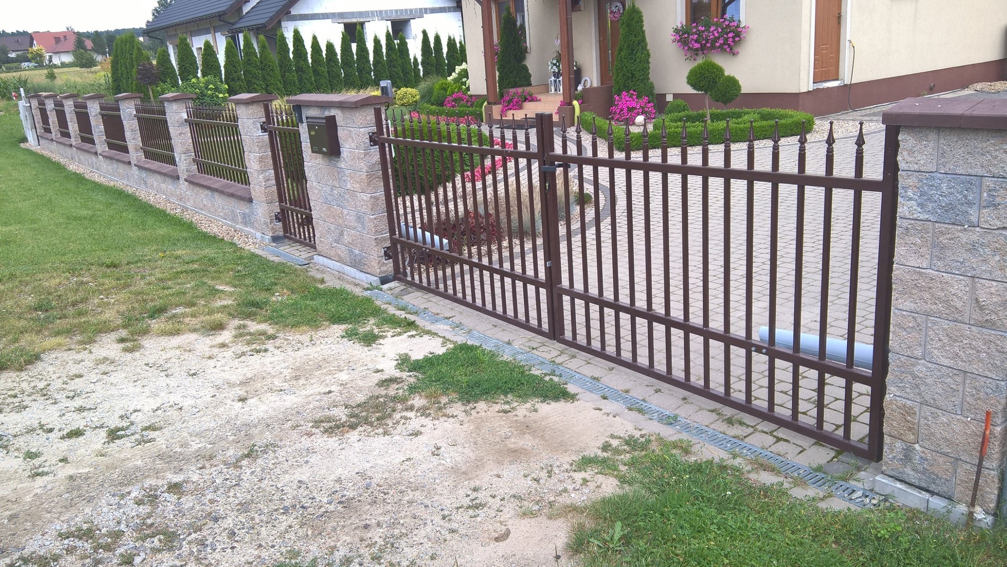 Nowoczesna brama wjazdowa z metalowymi prętami w kolorze brązowym, osadzona w kamiennych słupkach, z widocznym systemem automatycznego otwierania i zadbanym ogrodem w tle.