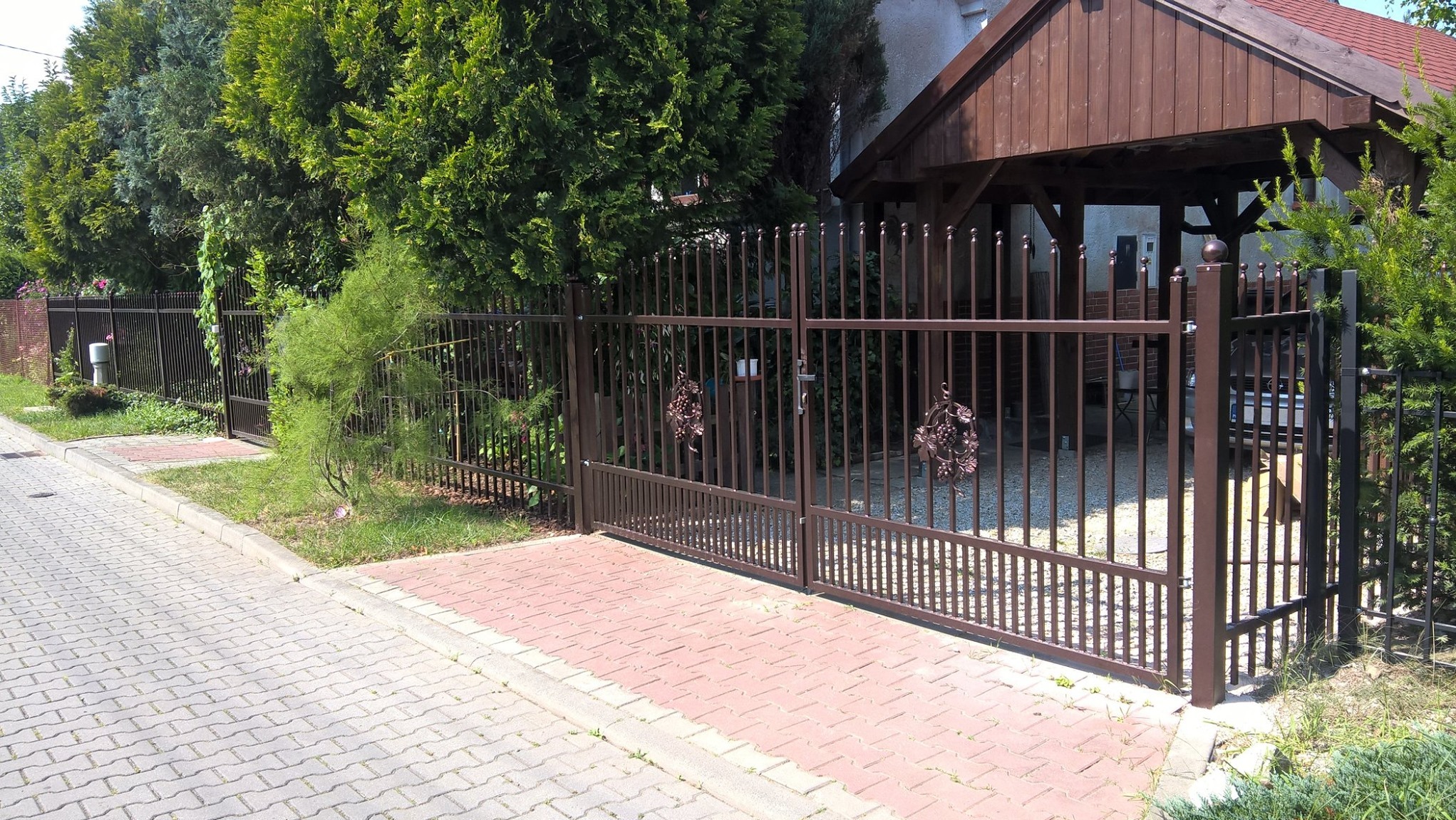 Brązowa metalowa brama wjazdowa z dekoracyjnymi elementami roślinnymi, prowadząca do posesji z wiatą garażową w tle.