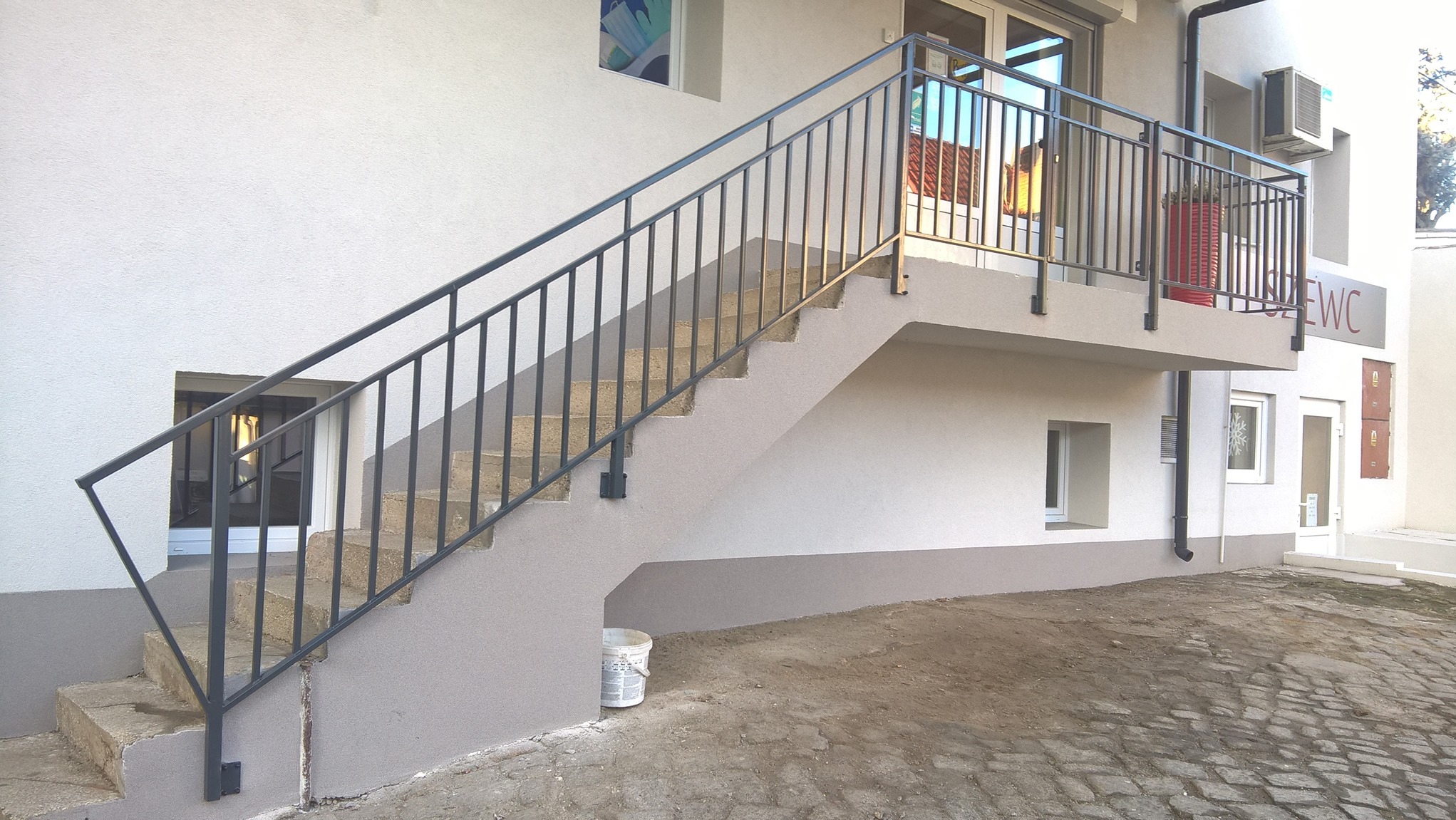 Zewnętrzne schody betonowe z prostą, metalową poręczą w kolorze grafitowym, prowadzące na mały balkon z metalową balustradą, widok z dołu, obok ściany budynku z małymi oknami.