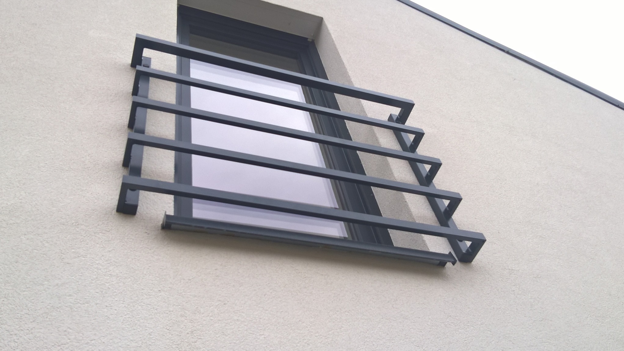 Nowoczesna, minimalistyczna krata okienna z szarych, prostokątnych profili na elewacji budynku o jasnej, chropowatej fakturze.