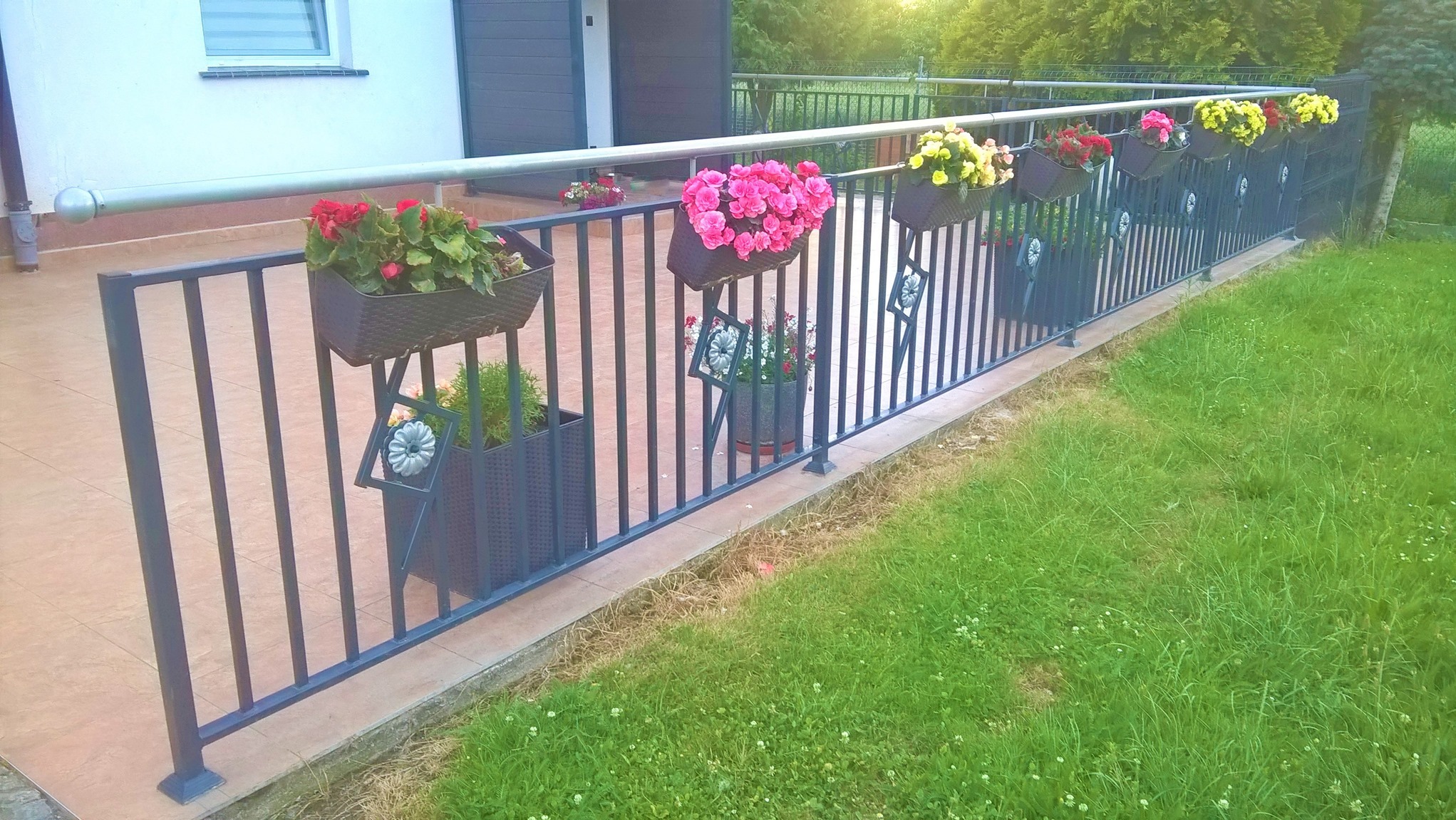 Ciemna, metalowa konstrukcja ogrodzenia z ozdobnymi elementami i wiszącymi donicami z kolorowymi kwiatami, widok z perspektywy trawnika.