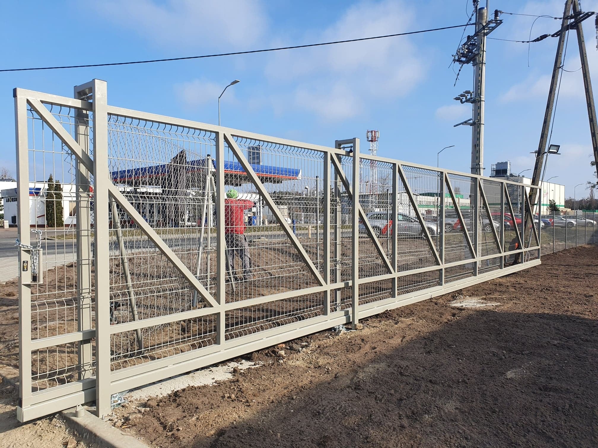 Długa, przesuwna brama przemysłowa z metalowej siatki, pomalowana na jasny kolor, z widocznym mechanizmem przesuwnym i betonową podstawą, na tle stacji benzynowej i słupów energetycznych.