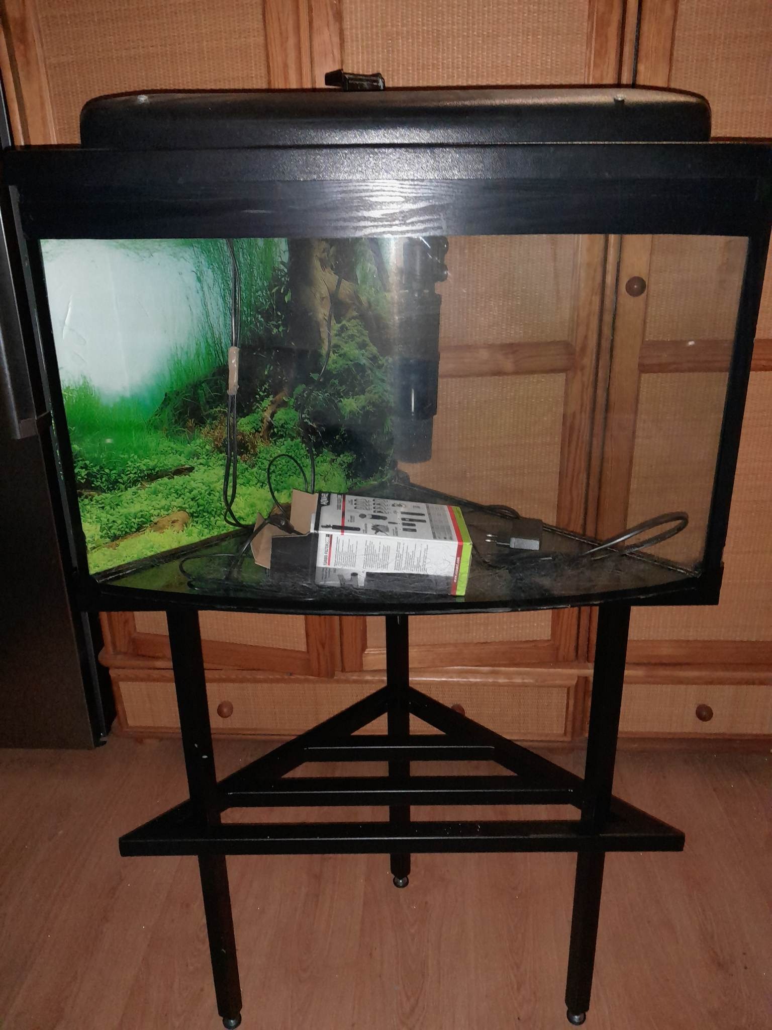 Czarny metalowy stelaż pod akwarium z trójkątną podstawą, na blacie akwarium filtr i akcesoria, tło z zieloną roślinnością wewnątrz akwarium.