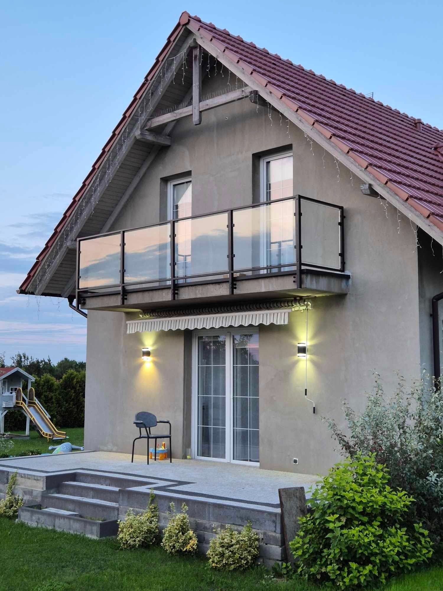 Dom z balkonem z czarną ramą i szklanymi panelami, oświetlony kinkietami, z tarasem i schodami wejściowymi, w tle plac zabaw dla dzieci.