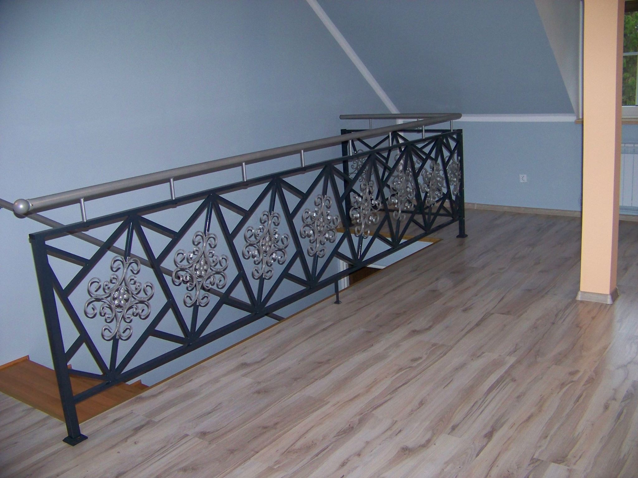 Kuta, czarna balustrada schodowa z ozdobnymi, srebrnymi rozetami i stalową poręczą, widoczna na tle błękitnej ściany i drewnianej podłogi.